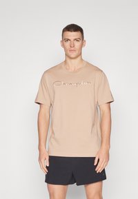 Beige Baumwoll-T-Shirt mit Rundhalsausschnitt, versehen mit aufgesticktem "Champion"-Logo in einer kontrastierenden Farbe auf der Vorderseite, kombiniert mit schwarzen Shorts.