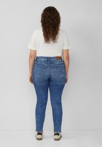 Kobieta z długimi kręconymi włosami, ubrana w białą koszulkę, niebieskie dżinsy i białe trampki, stoi twarzą do szarej ściany.