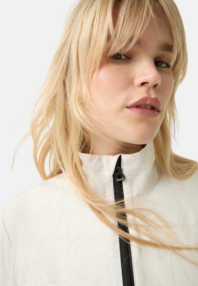 Weißer Reißverschlussjacke mit strukturiertem Muster, hohem Kragen und schwarzem Reißverschluss. Das Modell hat lange blonde Haare und einen neutralen Gesichtsausdruck.