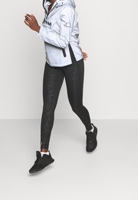 Jasnosrebrna wiatrówka z logo na przodzie i bocznymi rozcięciami, w zestawie z czarnymi legginsami w wzór węża i czarnymi trampkami.