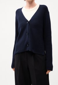 Tmavě modrý pletený kardigan s výstřihem do V, žebrovanou strukturou, čtyřmi černými knoflíky a mírně zkráceným střihem. Kombinovaný s krémovým topem a černými kalhotami.