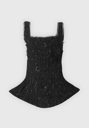 Femme portant un top noir texturé sans manches à basque et un pantalon large bleu marine avec des chaussures blanches pointues, posant avec une main sur la hanche.