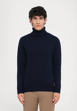 Suéter azul marino de cuello alto en textura acanalada con mangas largas y silueta ajustada, combinado con chinos de color marrón claro.