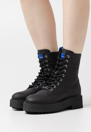 Black leather ankle boots with a textured finish, round toe, and thick, ridged sole. Features silver eyelets and blue logo accents on the tongue.