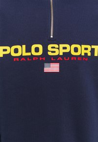 Dunkelblaues Kleidungsstück mit gelbem "POLO SPORT"-Text, rotem "RALPH LAUREN"-Text, kleiner US-Flagge und einem silbernen Reißverschluss über dem Text.