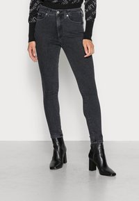 Jeans negros ajustados con un ligero brillo, cintura alta, bolsillos frontales y costuras en contraste, combinados con botines negros de tacón.