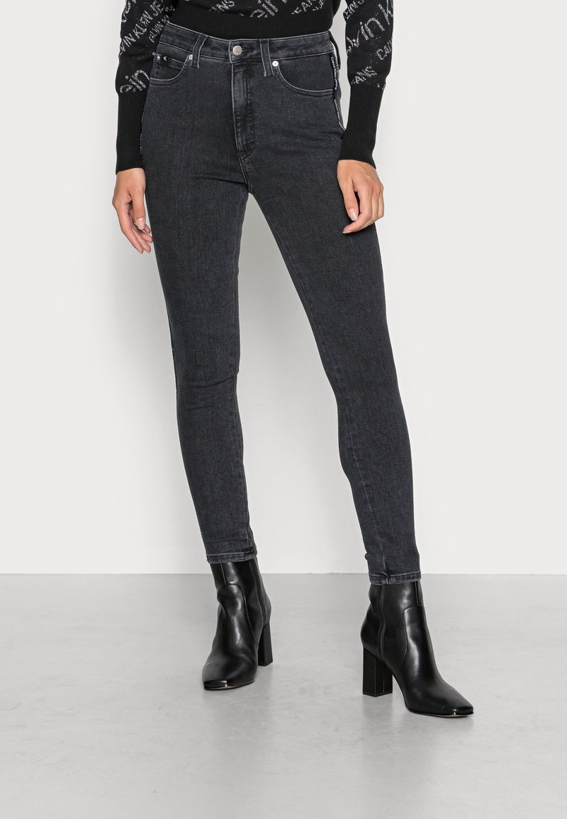 Jeans negros ajustados con un ligero brillo, cintura alta, bolsillos frontales y costuras en contraste, combinados con botines negros de tacón.