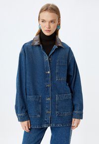 Jeansjacka med en krage i brunt läder, framsknappa, två bröstfickor och en avslappnad passform. Mörkblå färg med subtil textur.