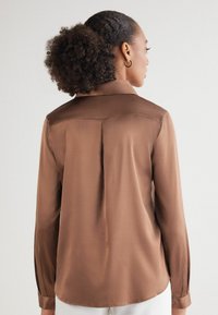 Bruine satijnen shirt met een kraag, lange mouwen met knoopmanchetten, en een licht gebogen zoom met een plooi aan de achterkant.