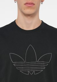 Melna kokvilnas T-kreklā ar lielu logo dizainu, kas izveidots no sudraba rhinestones, stilizētas lapas un horizontālām līnijām.