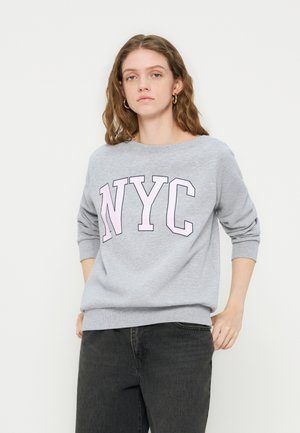 Young woman with curly hair wearing gold hoop earrings, a grey sweatshirt with "NYC" lettering, and black jeans, standing with hands in pockets.