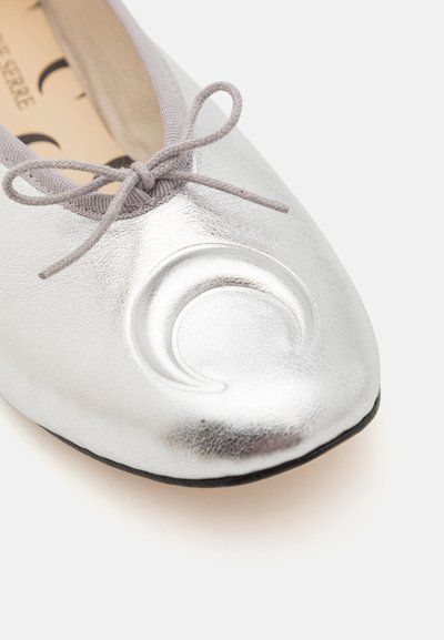 Silver metallic flat shoe with a rounded toe, featuring a textured finish and a gray satin bow. Notable design accent includes a circular motif.