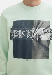 Felpa verde menta con stampa grafica in bianco e nero, con il testo "DENIM TOM TAILOR," e una texture liscia.