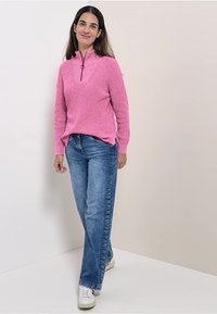 Jasnoróżowy sweter dzianinowy z kołnierzem na pół-zamek, zestawiony z prostymi niebieskimi dżinsami i białymi trampkami, stojący na neutralnym tle.