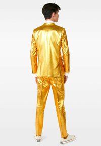 Completo in satin oro con giacca aderente e pantaloni a sigaretta, caratterizzato da una texture lucida. Indossato con una camicia bianca e sneakers bianche casual.