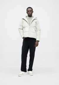 Giacca imbottita color crema con cappuccio, tasche frontali e chiusura con zip, abbinata a pantaloni scuri e scarpe bianche. Tessuto morbido.
