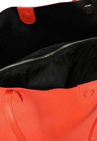 Grote oranje leren totebag met een gladde textuur, zwart binnenvak en een zilveren ritssluiting. Heeft een rond accent aan de rand.