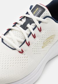 White athletic shoe with mesh upper, featuring red and navy accents, laces, and a textured sole. Logo displayed on the side and tongue.