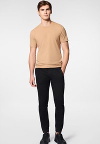 Beżowa koszulka z krótkim rękawem w połączeniu z czarnymi dopasowanymi spodniami. Spodnie mają mankiety na kostkach, a model ma na sobie czarne trampki. Gładka struktura materiału.