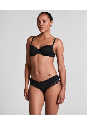 Kobieta stoi, ubrana w czarny biustonosz z koronkową lamówką i dopasowane majtki, z włosami związanymi do tyłu i neutralnym tłem.