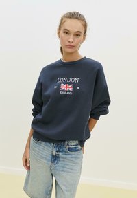 Námornícky modrý sveter s vyšitým nápisom "LONDÝN" a dizajnom britskej vlajky. Voľný strih s rebrovanými nápletmi a klasickým okrúhlym výstrihom. Nosí sa s svetlomodrými džínsovými šortkami.