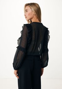 Blusa nera trasparente con pois, dettagli arricciati su maniche e spalle, design corto e maniche lunghe a sbuffo, abbinata a pantaloni neri.