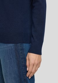 Marineblauer Strickpullover mit gerippten Bündchen, kombiniert mit Jeans. Die Textur wirkt weich mit einem taillierten Schnitt, Hand an der Seite gezeigt.
