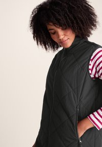 Woman with curly hair wearing a black quilted vest over a striped long-sleeve shirt, standing with one hand in vest pocket.