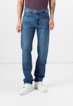 Mand iført blå jeans med lige ben, hvide sneakers og en mørkelilla skjorte, står foran en ensfarvet hvid baggrund.