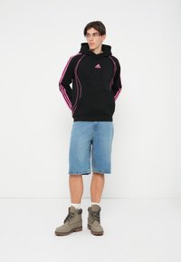 Čierna mikina s kapucňou a ružovými akcentmi, na ktorej je logo Adidas, skombinovaná s modrými denimovými šortkami a šedými topánkami. Mikina má kapucňu so šnúrkami.