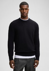 Schwarzer Pullover aus einem strukturierten, flauschigen Stoff mit Rundhalsausschnitt sowie gerippten Bündchen und Saum, getragen über einem weißen Hemd. Formelle graue gemusterte Hose.