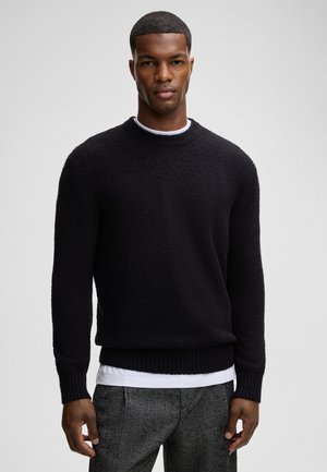 Zwarte sweater van een textuur met een zachte stof, met een ronde halslijn en geribbelde manchetten en zoom, gedragen over een witte blouse. Formele grijze broeken met een patroon.