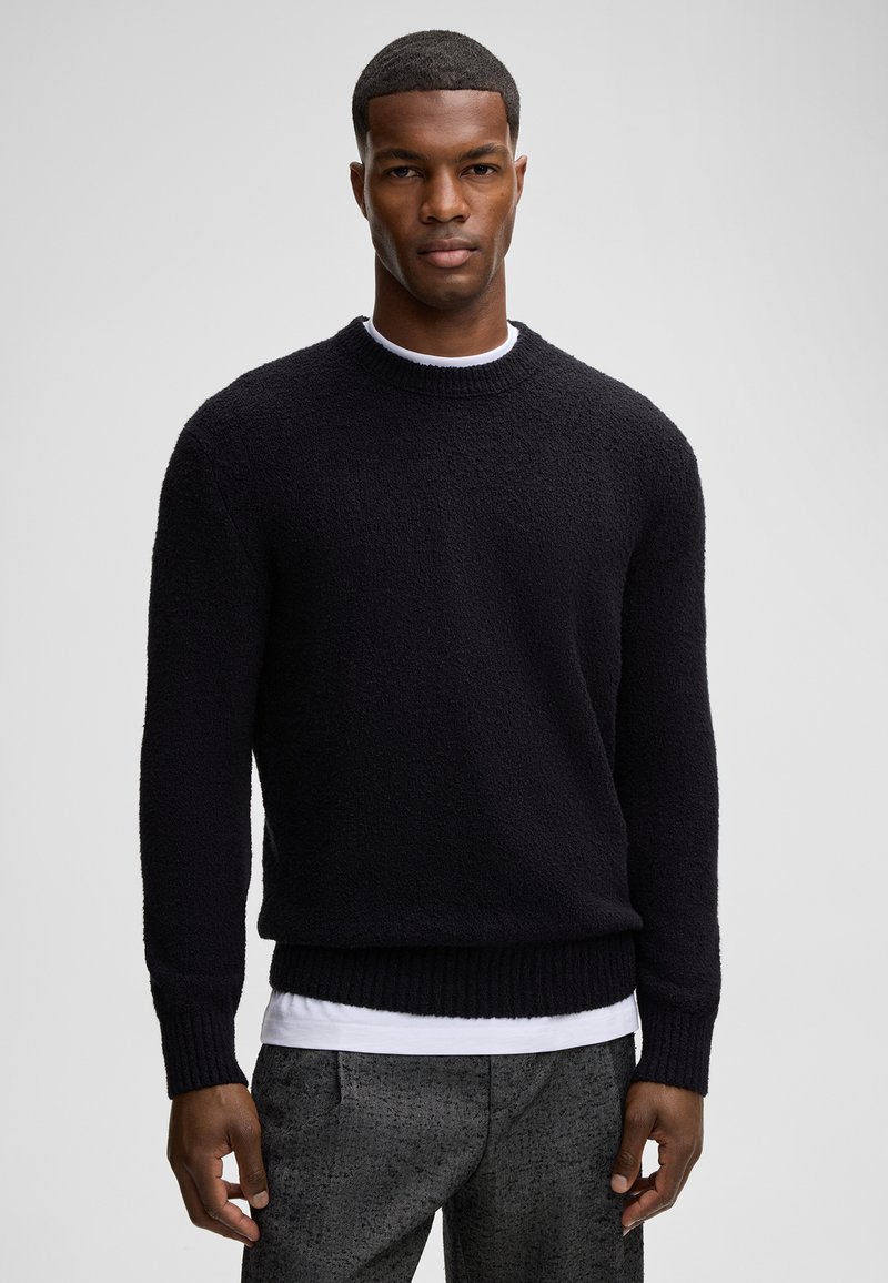 Schwarzer Pullover aus einem strukturierten, flauschigen Stoff mit Rundhalsausschnitt sowie gerippten Bündchen und Saum, getragen über einem weißen Hemd. Formelle graue gemusterte Hose.