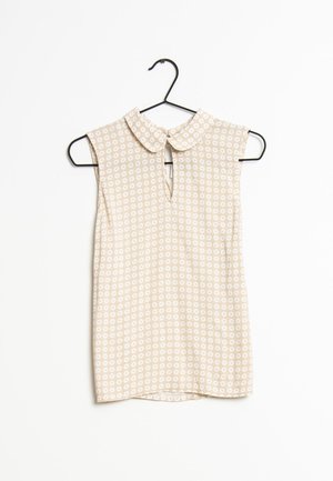 Blouse beige sans manches avec motif à pois blancs, col Claudine et fente avant, suspendue sur un cintre noir sur fond blanc.