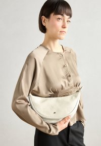 Pochette blanche à motifs avec une forme arrondie, tenue sous le bras. Présente une texture lisse, des éléments en or, et une fermeture éclair.