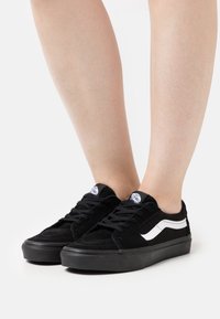 Baskets en daim noir avec une bande latérale blanche, une semelle en caoutchouc plate et des lacets noirs. Le design présente un bout rond et un marquage minimal.