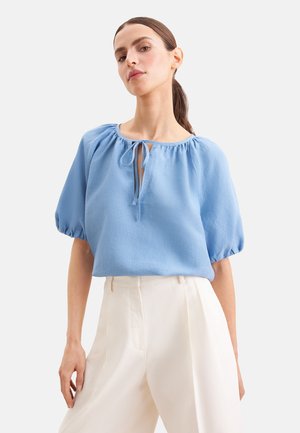 Vrouw poseert in een lichtblauw bloesje met pofmouwen, keyhole-hals met strik en witte, hoog getailleerde wijde broek.