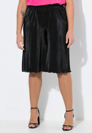 Zwarte geplooide culottes met een geschulpte zoom; gemaakt van een gestructureerde stof. Gecombineerd met zwarte sandaaltjes met bandjes en een roze top.