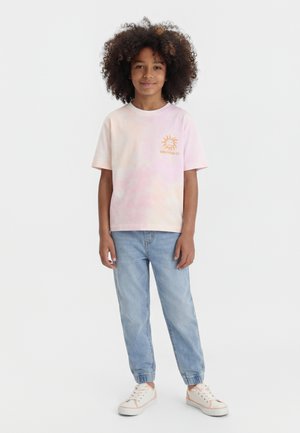 Barn med krøllet hår iført en lyserød tie-dye t-shirt med solmotiv, lyseblå jogger-jeans og hvide sneakers med lyserøde detaljer.