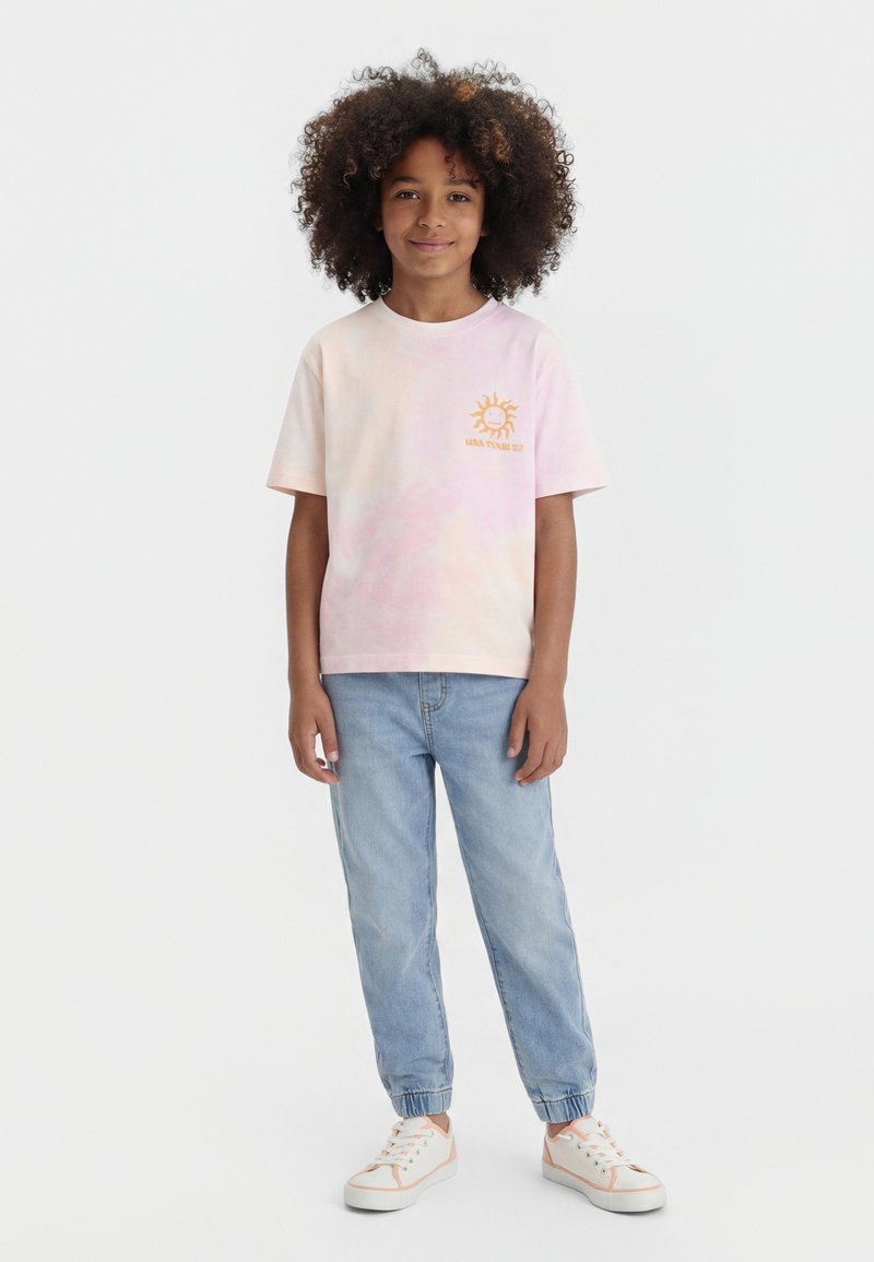 Dijete kovrčave kose koje nosi svijetloružičastu tie-dye majicu sa suncem, svijetloplave traperice jogger kroja i bijele tenisice s ružičastim detaljima.