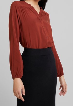 Kobieta ubrana w długą, ceglastoczerwoną koszulę zapinaną na guziki, wpuszczoną w wysokowaistową czarną spódnicę na tle gładkiego szarego tła.