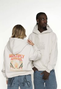 Bílé mikiny s tlumenými grafickými potisky. Jedna má výrazný nápis "MULTIPLY APPAREL" v sytě červené barvě, druhá má jemně vyšívané logo. Viditelné džínové kalhoty.