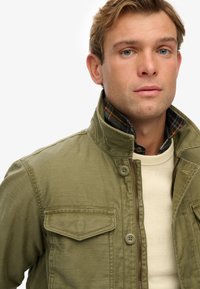 Ung mand med blå øjne, iført en lagdelt khaki-jakke over en cremefarvet sweater, mod en hvid baggrund.