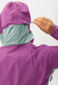 Veste imperméable violette et bleu clair avec capuche, dotée d'un cordon de serrage et d'un tissu texturé. Détail de poche zippée sur le côté.