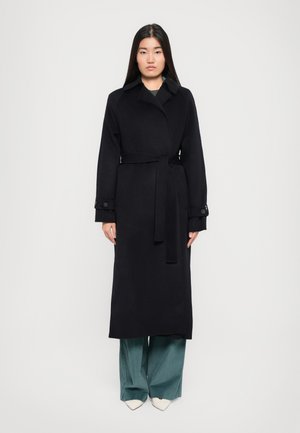 Black wool trench coat with a belt, large collar, and buttoned cuffs. Worn over teal wide-leg pants and white pointed-toe shoes.