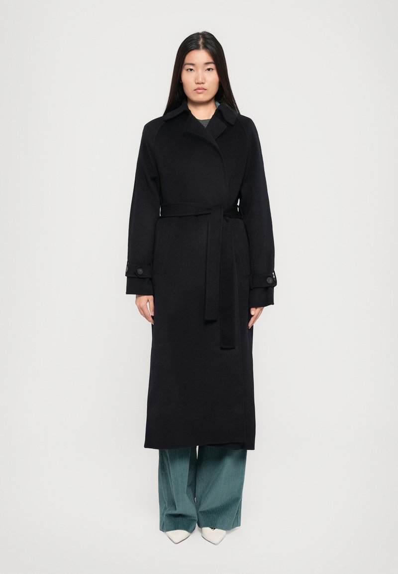 Abrigo de lana negro con cinturón, gran cuello y puños abotonados. Lleva puesto sobre pantalones de pierna ancha en color azul verdoso y zapatos blancos de punta.