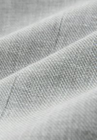 Hellgrauer Stoff mit einer strukturierten Webart, der feine Muster und weiche, glatte Oberflächen aufweist. Nahaufnahme mit dezenten Nähdetails.