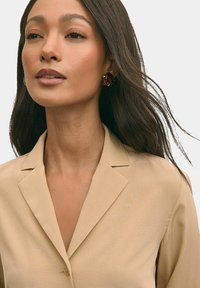 Blazer beige chiaro con colletto a reversal, chiusura a un bottone e texture liscia. Il modello sfoggia un'acconciatura elegante e accessori ridotti al minimo.