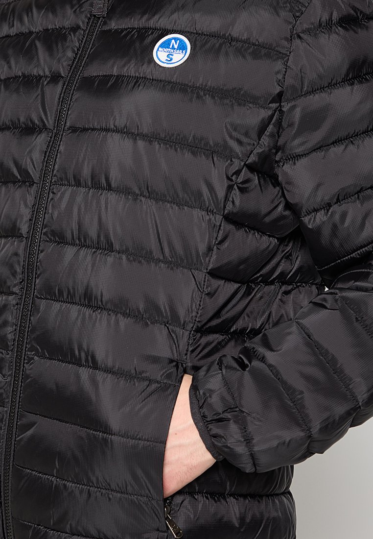 Giacca a vento nera con sezioni a cucitura verticale, zip frontale, polsini elasticizzati e un logo circolare sul petto. Texture liscia.