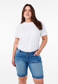 Camiseta blanca de algodón combinada con pantalones cortos de mezclilla azul. Los pantalones cortos tienen un diseño de cinco bolsillos, cierre frontal con botón y dobladillo vuelto.